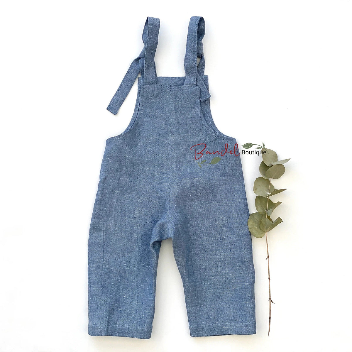 A pair of baby denim overall shorts in a medium blue chambray fabric, laid flat on a white background next to a green eucalyptus sprig. A small embroidered logo on the chest reads 'Buncha Boutique'.