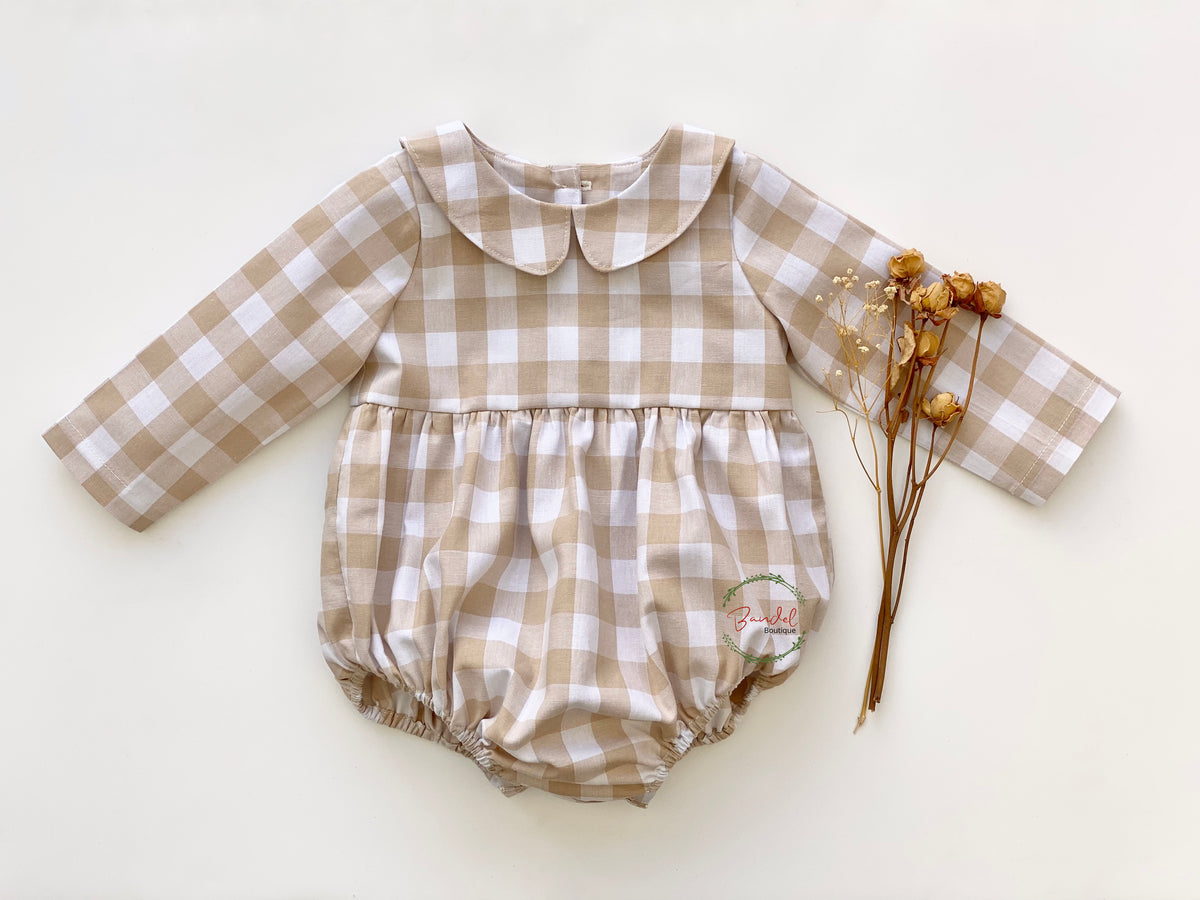 A beige check romper with long sleeves, a Peter Pan collar, elastic at the leg, and wooden buttons at the back, displayed on a flat surface.