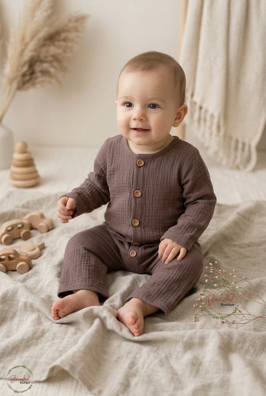 A baby sitting on a beige linen blanket wearing a matching short-sleeve button-front romper and pants in a muted taupe-brown textured fabric. The scene is styled with wooden toys, dried grasses, and a cream knit throw in a soft indoor setting.