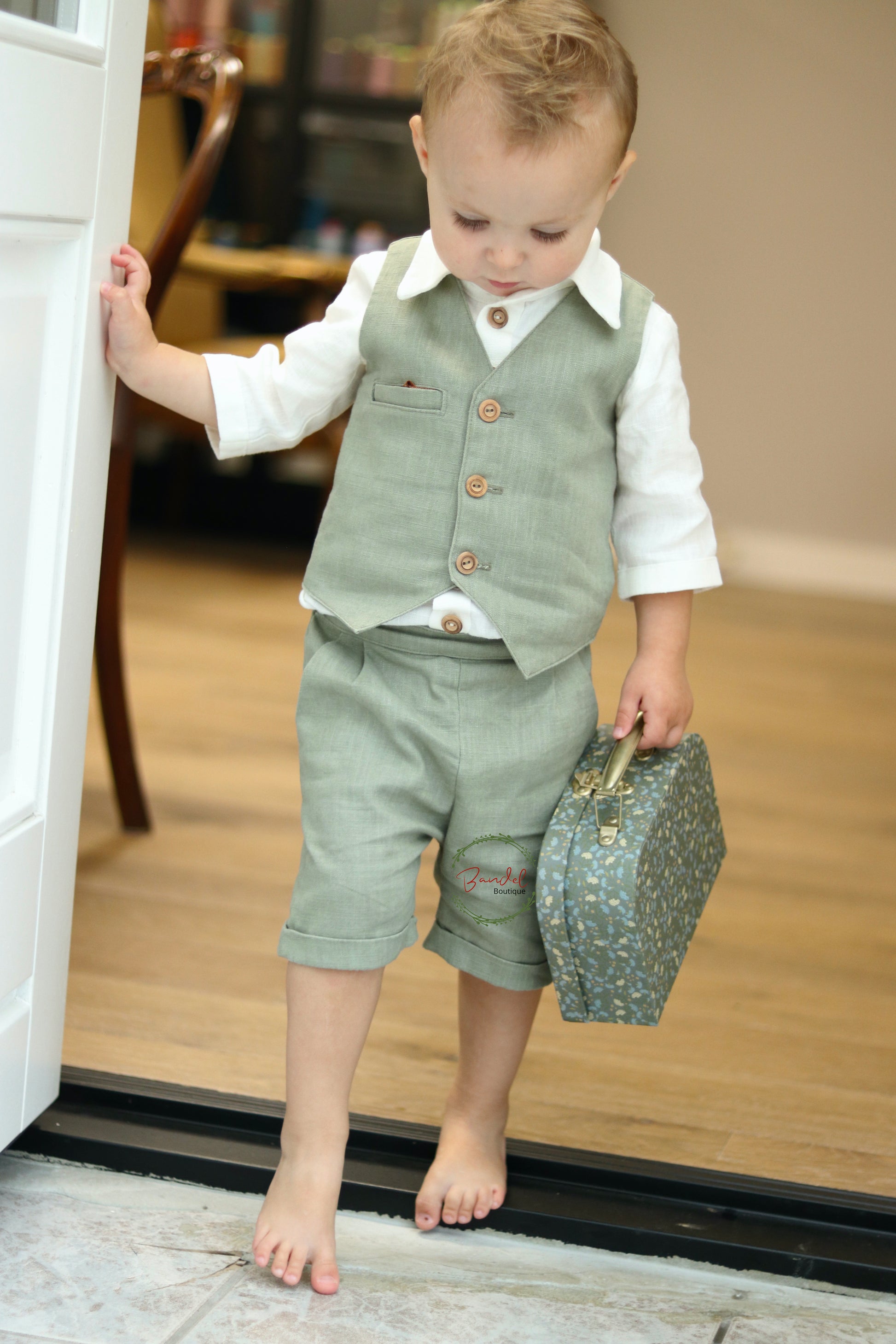 A young child wearing a dark-mint green 3-piece short pageboy outfit with a vest and a collared shirt, holding a blue gift box.