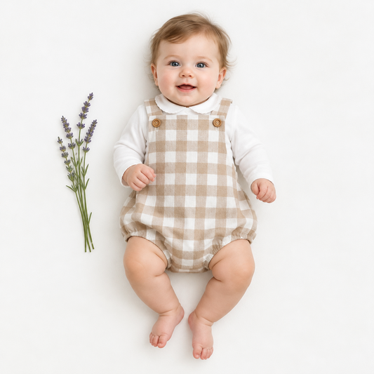 A baby lying on a white background wearing a white long-sleeve shirt and a beige-and-white gingham romper with brown buttons. A small sprig of lavender is placed to the left of the baby.