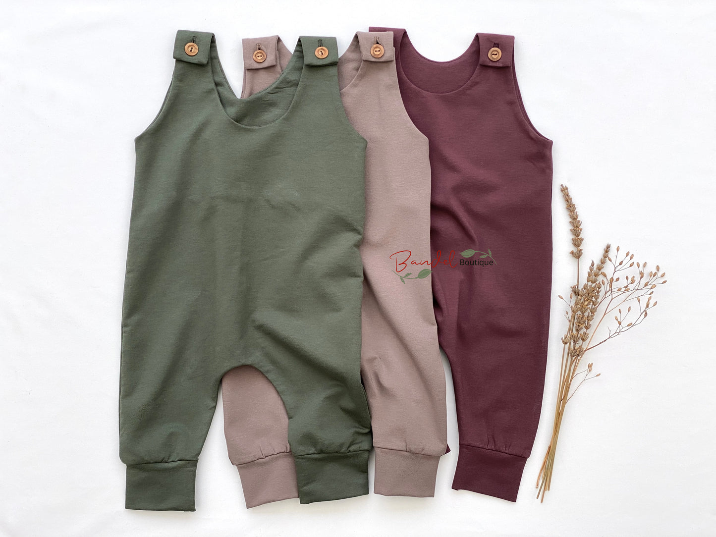 Three children's rompers in green, beige, and purple on a white background with dried plants.