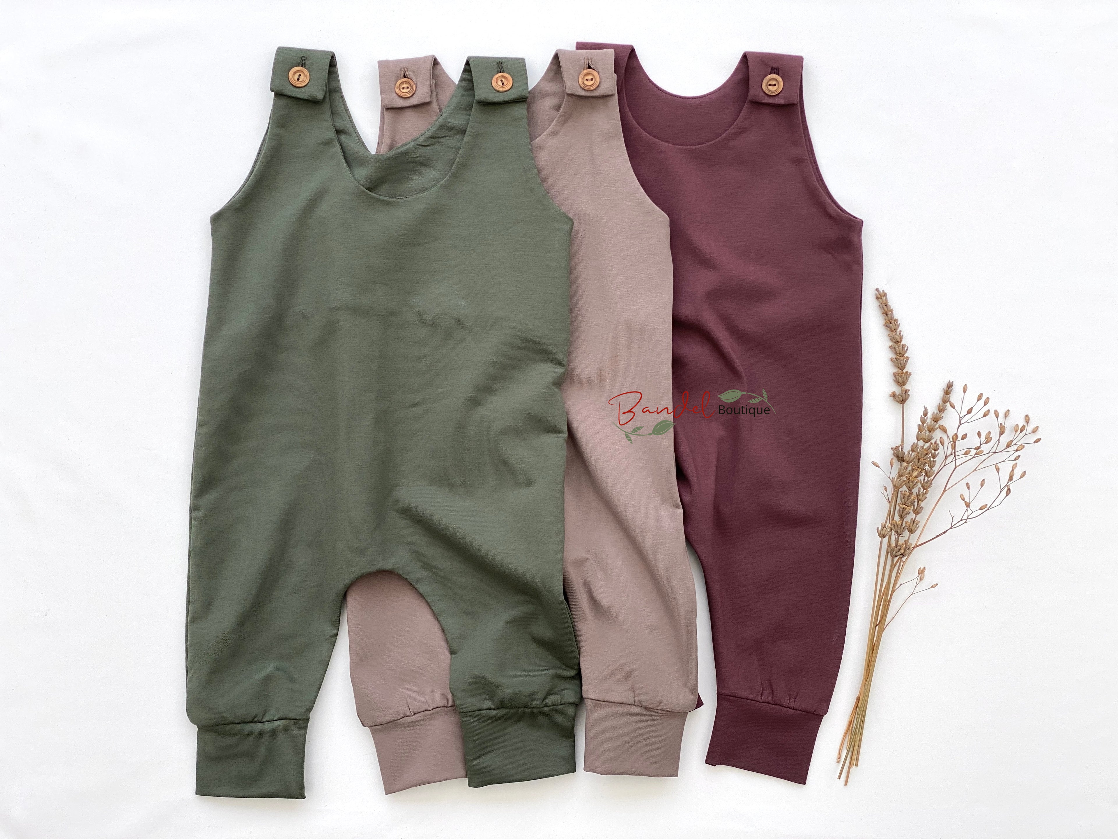Three children's rompers in green, beige, and purple on a white background with dried plants.