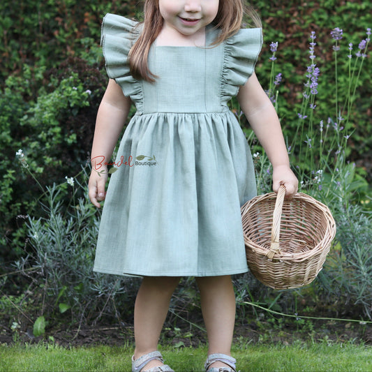 A young girl with brown hair in a ponytail stands in a garden wearing a sage green dress and holding a small wicker basket.