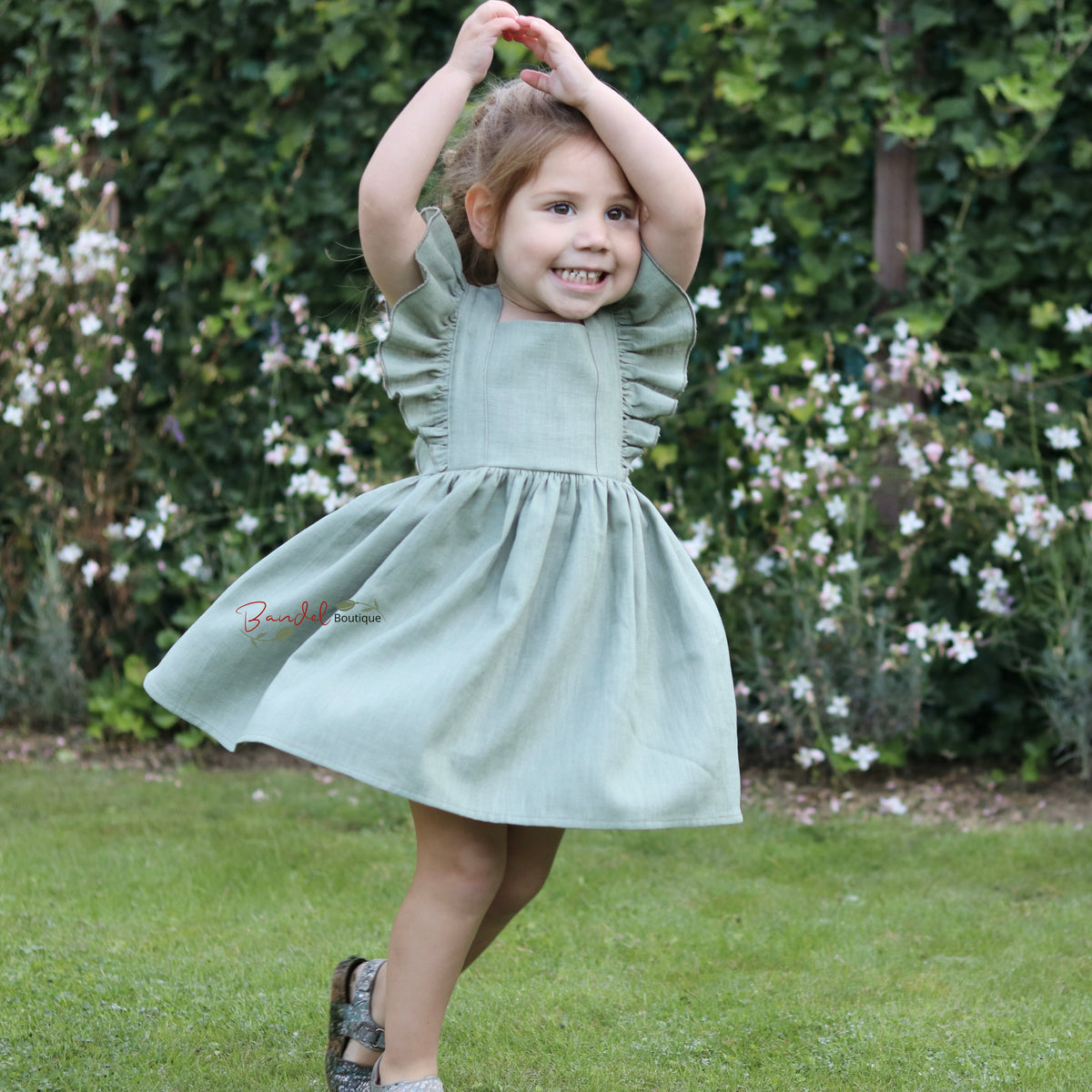 A young girl with brown hair smiles and twirls outdoors in a light sage green dress, standing on a green lawn with a leafy hedge and white flowers in the background.