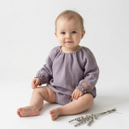 A baby sitting on a white studio floor wearing a dusty purple long-sleeve gauze romper, with a small tan brand patch on the sleeve and dried lavender sprigs nearby.