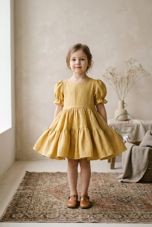 A young girl stands on a patterned rug in a softly lit room, wearing a mustard yellow tiered dress with puff sleeves and brown shoes. A vase with dried stems and a draped throw are visible in the background.