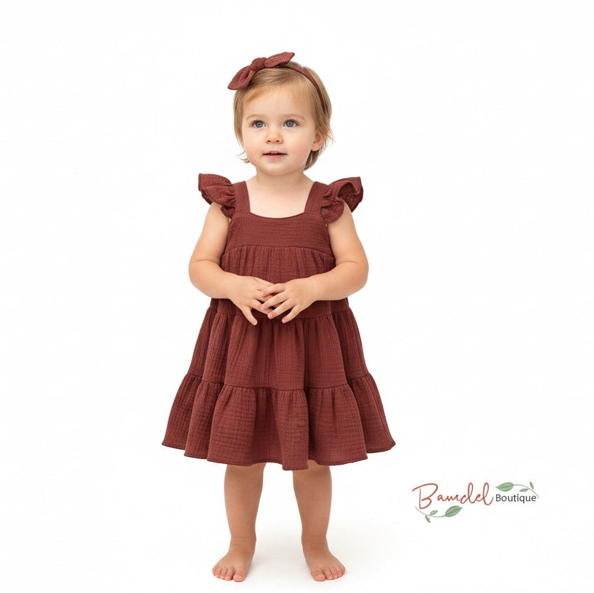 Young girl wearing a brown dress with ruffled sleeves and a bow on her head against a white background.