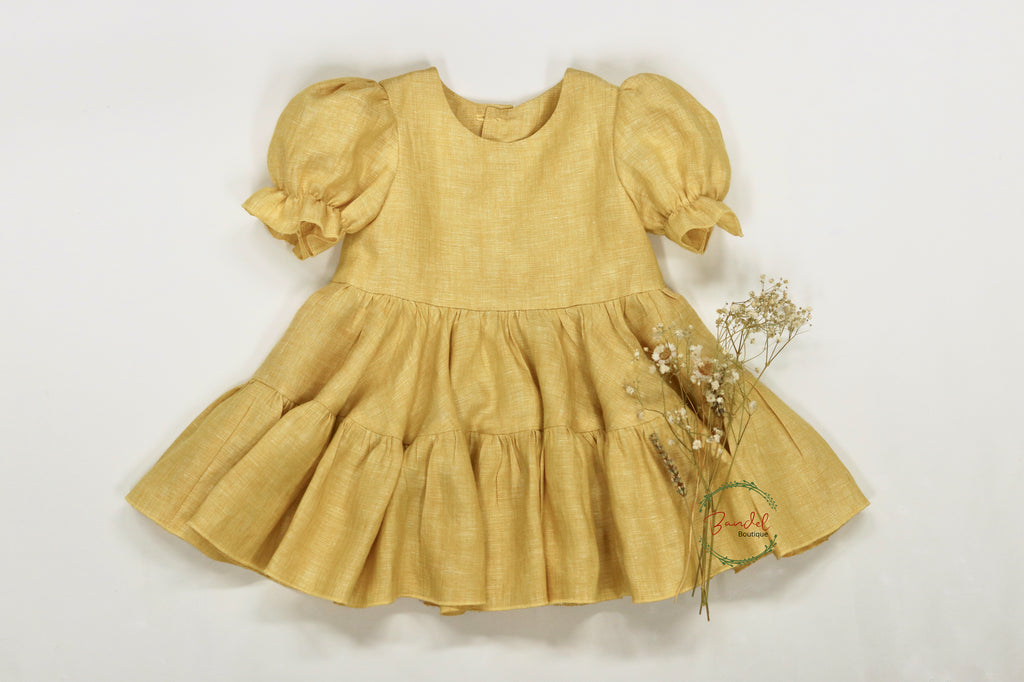 A yellow linen dress with short puff sleeves and a two-tiered gathered skirt, displayed with a wooden button back closure and some dried flowers for decoration.