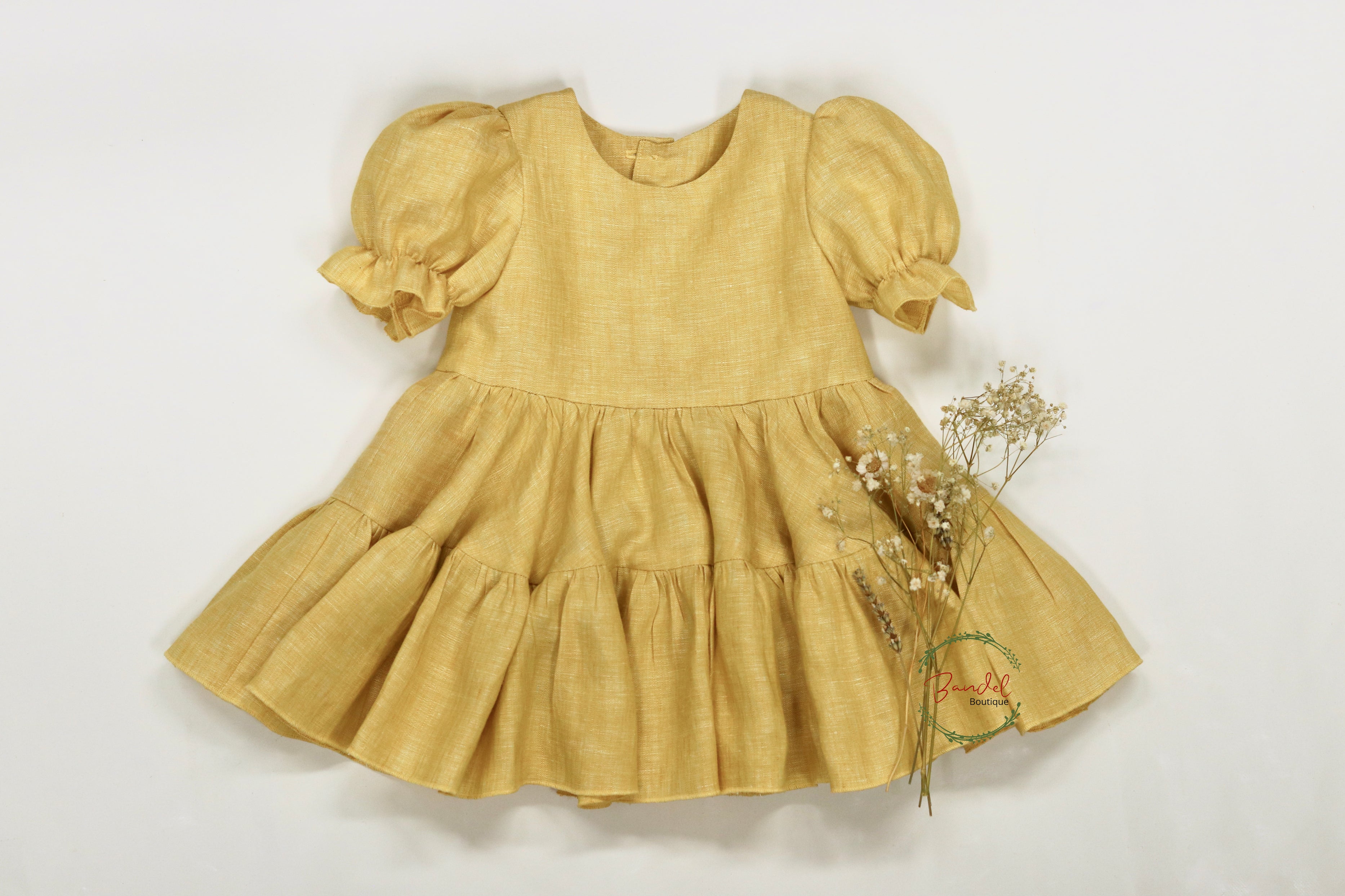 A yellow linen dress with short puff sleeves and a two-tiered gathered skirt, displayed with a wooden button back closure and some dried flowers for decoration.