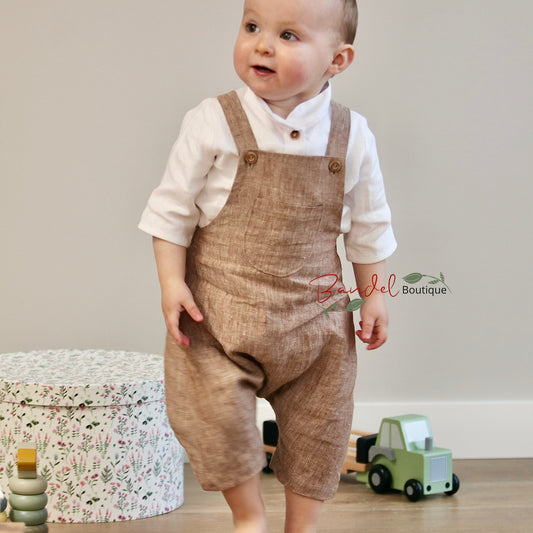 Baby wearing brown overalls and a white shirt in a room with toys and a floral-patterned box.