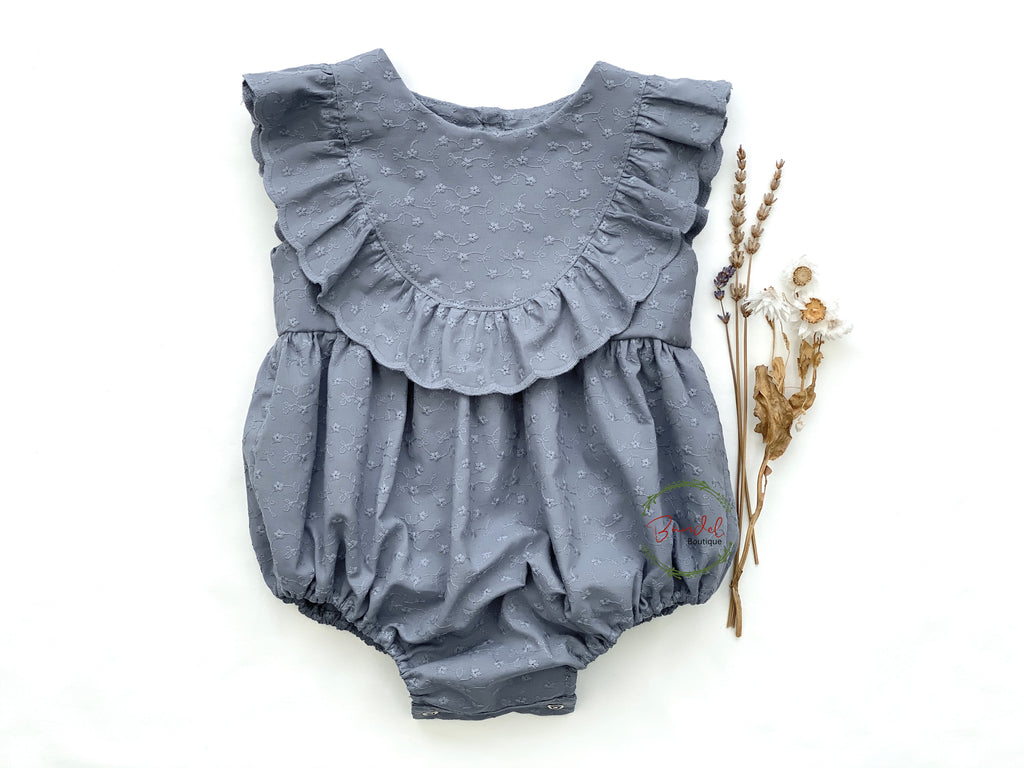 A dusty-blue romper with ruffled front bodice and embroidery details, displayed with wooden button back closure and a small wooden object for scale reference.