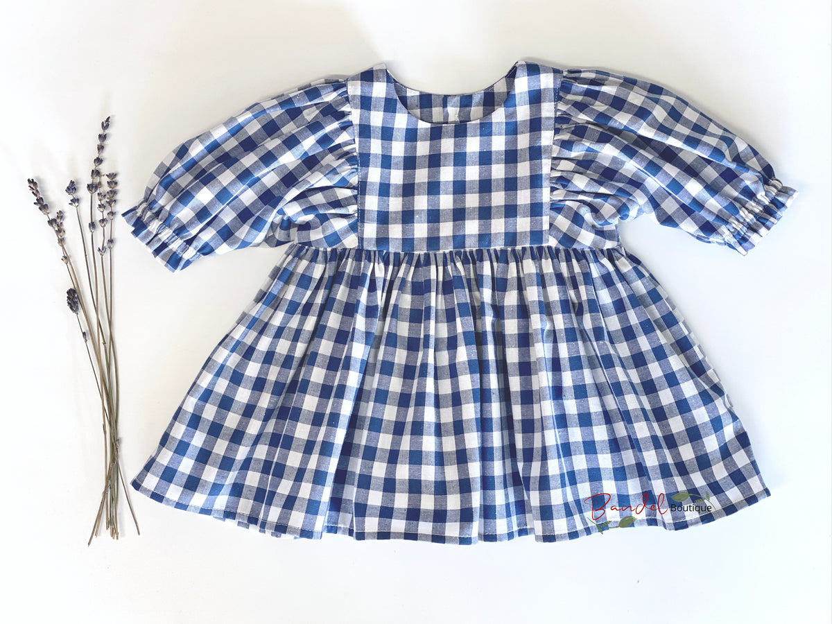 A blue and white gingham baby dress with puff sleeves and a gathered skirt, laid flat on a white background next to dried lavender sprigs.