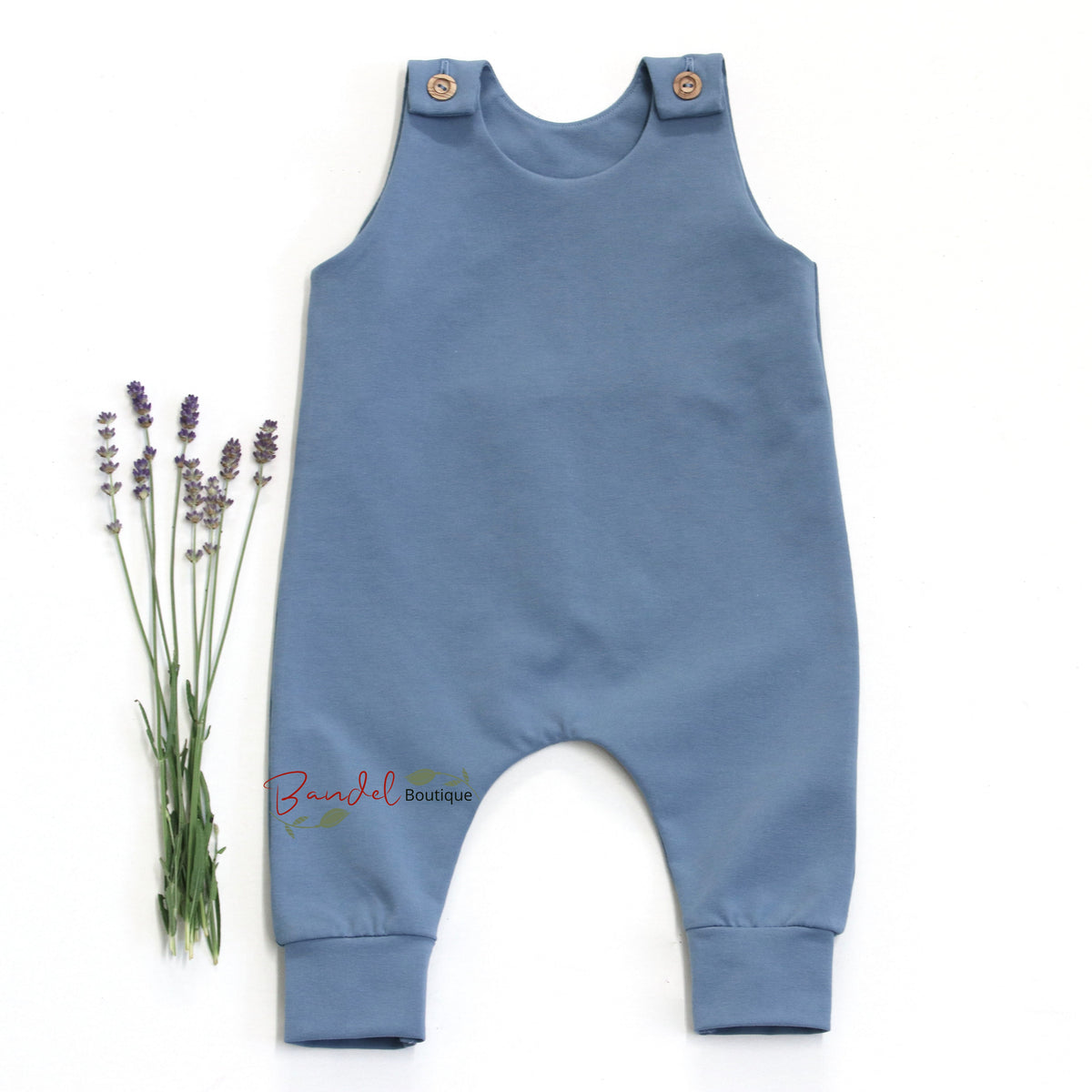 A dusty blue baby romper with snap shoulder straps and a small embroidered 'Jodelt linen' logo near the leg, displayed flat on a white background next to a small bunch of lavender.