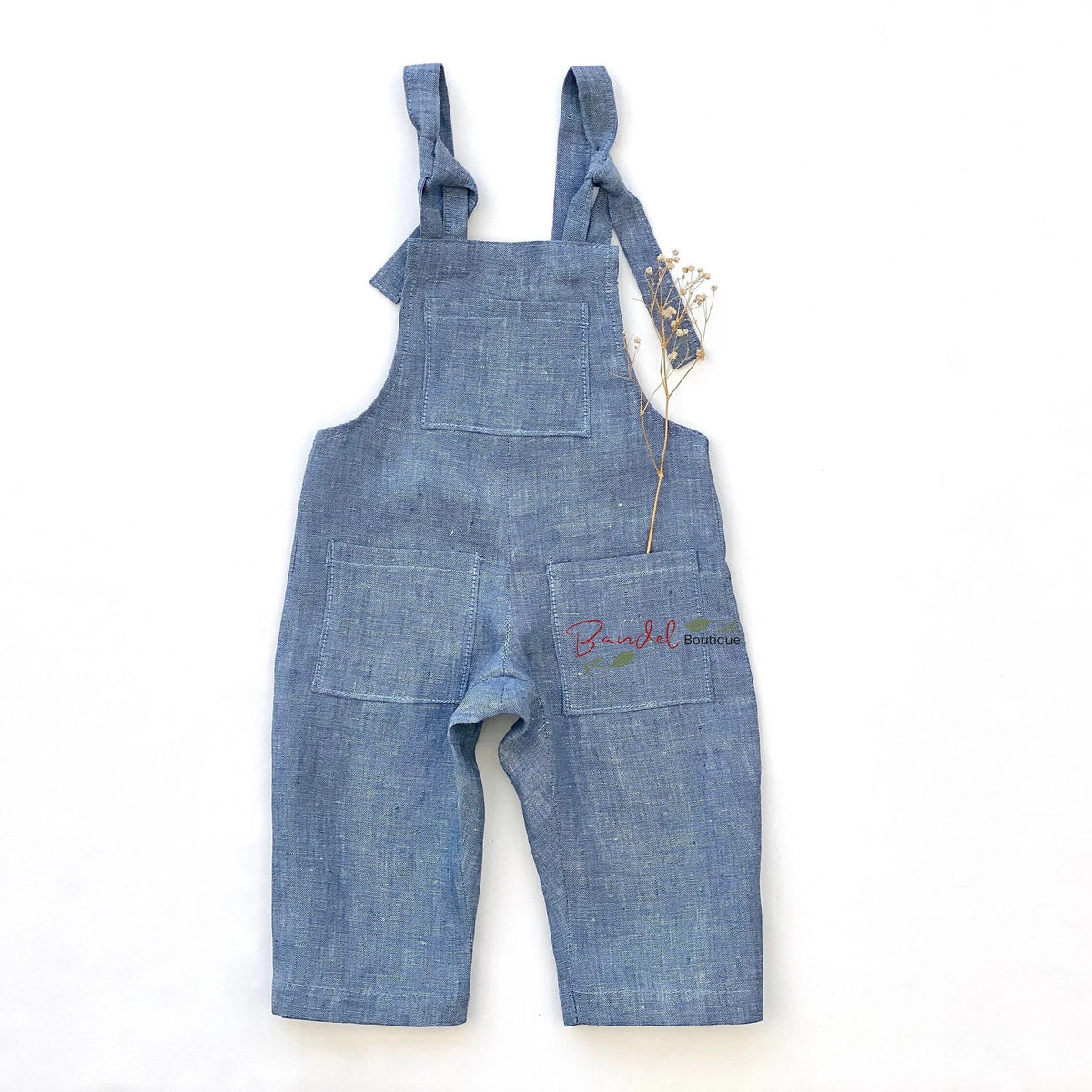 A pair of blue denim baby overalls with adjustable shoulder straps, laid flat on a white background. The overalls feature a front bib pocket, two front patch pockets, and a small colorful embroidered logo on the right pocket.