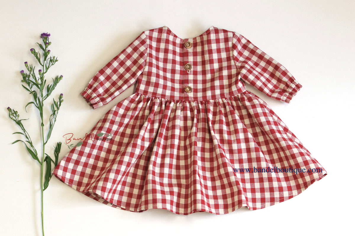 A red and white gingham long-sleeve dress for toddlers, featuring a buttoned neckline and a flared skirt, laid flat on a white background next to a green plant stem.
