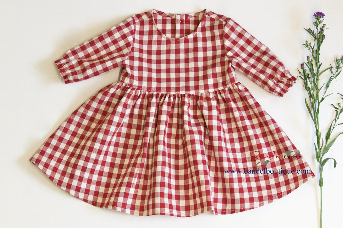 A red and beige gingham check long-sleeve dress for toddlers, laid flat on a white background with a sprig of greenery on the right side.