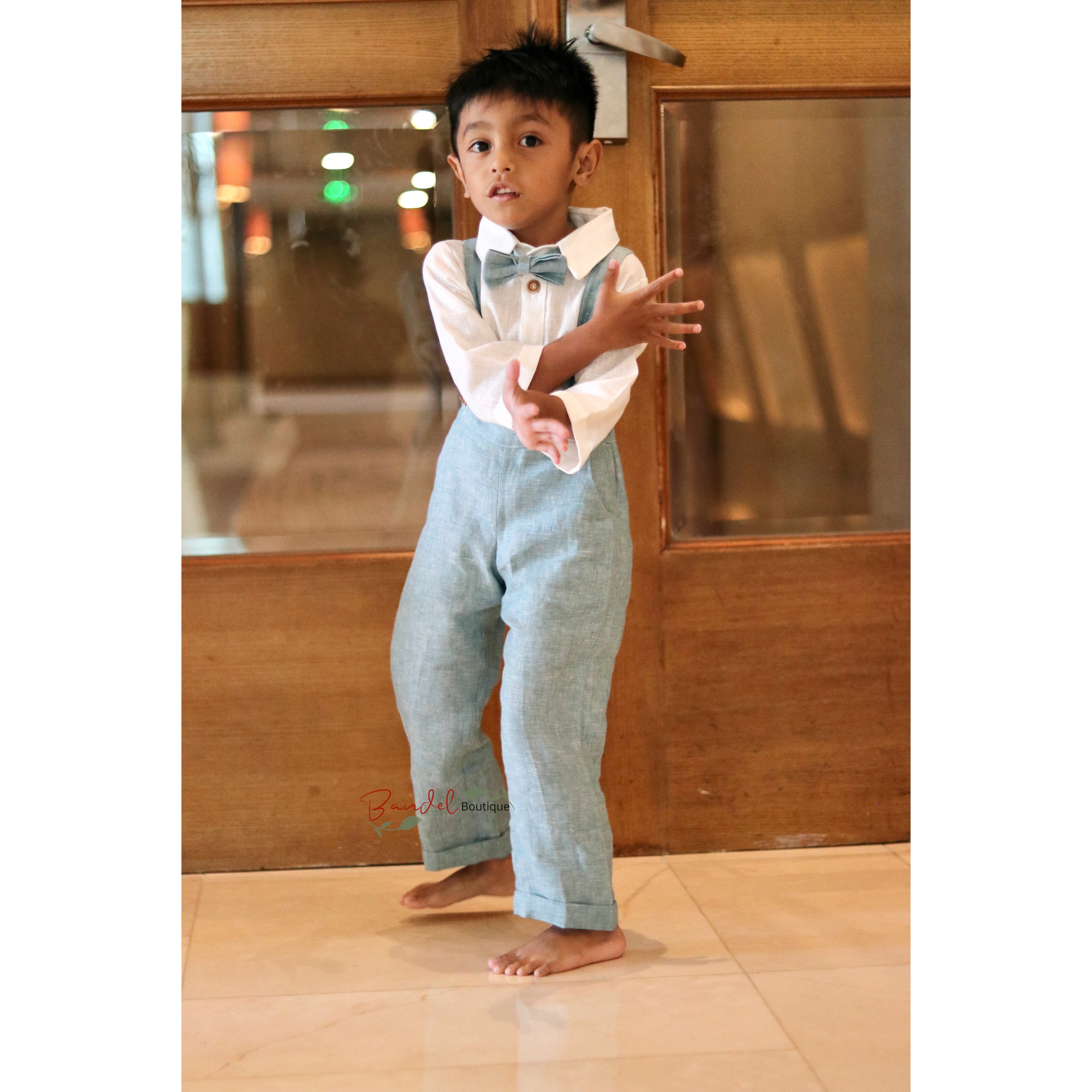 Child wearing a white shirt, blue bow tie, and light blue pants standing in front of a wooden door.