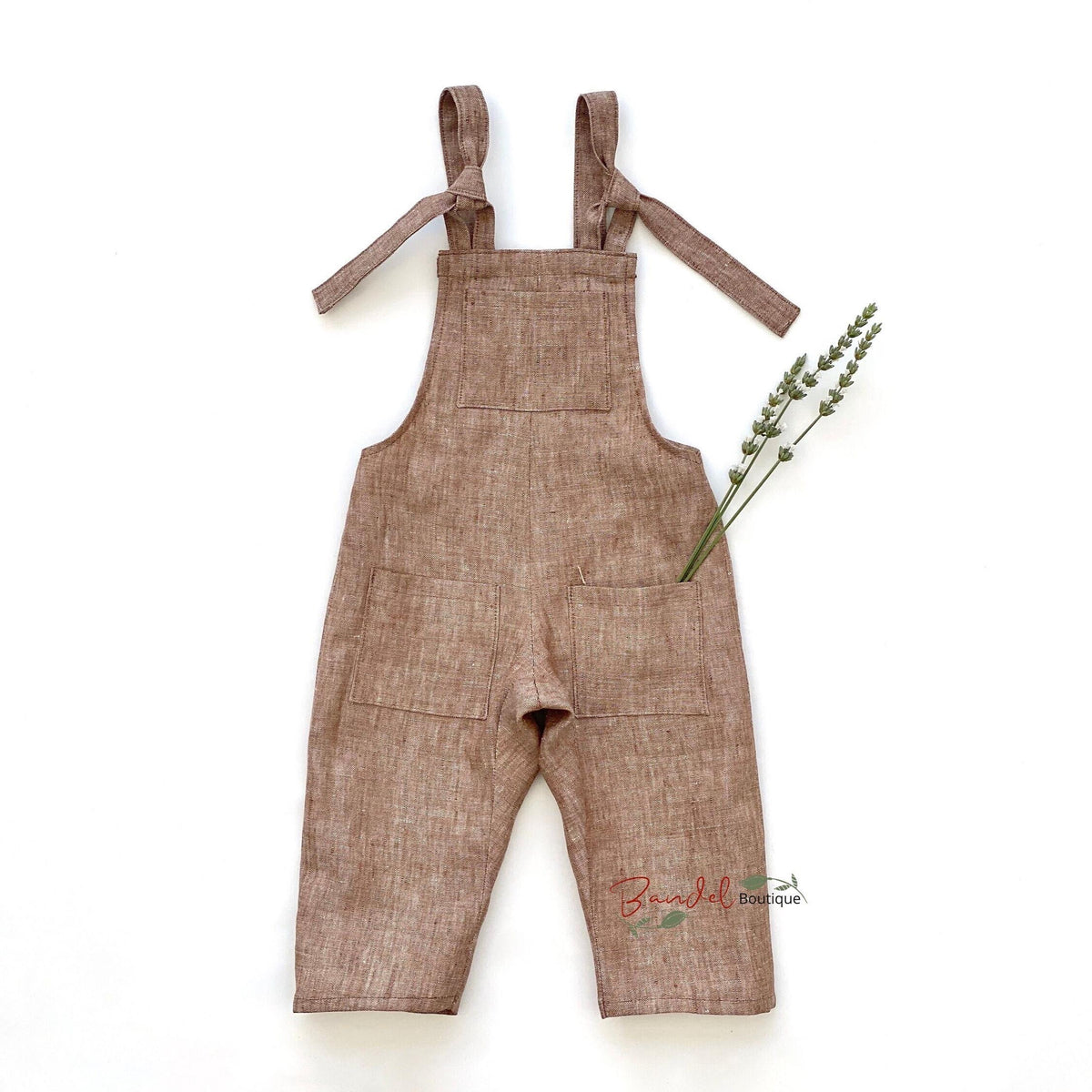 A baby romper in a textured brown linen-like fabric, featuring tie-shoulder straps, a bib-style chest panel, and front pockets, laid flat on a white background with a sprig of lavender.
