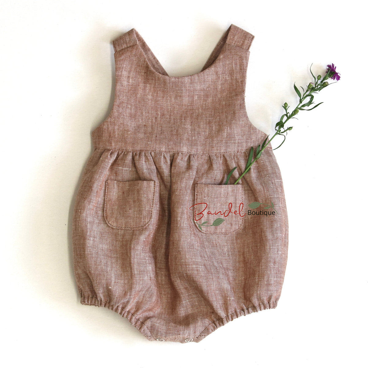 A sleeveless baby romper in a textured, dusty rose-brown linen-like fabric, featuring a front pocket with 'Brunchel Boutique' embroidered on it, styled with a small purple flower on a white background.