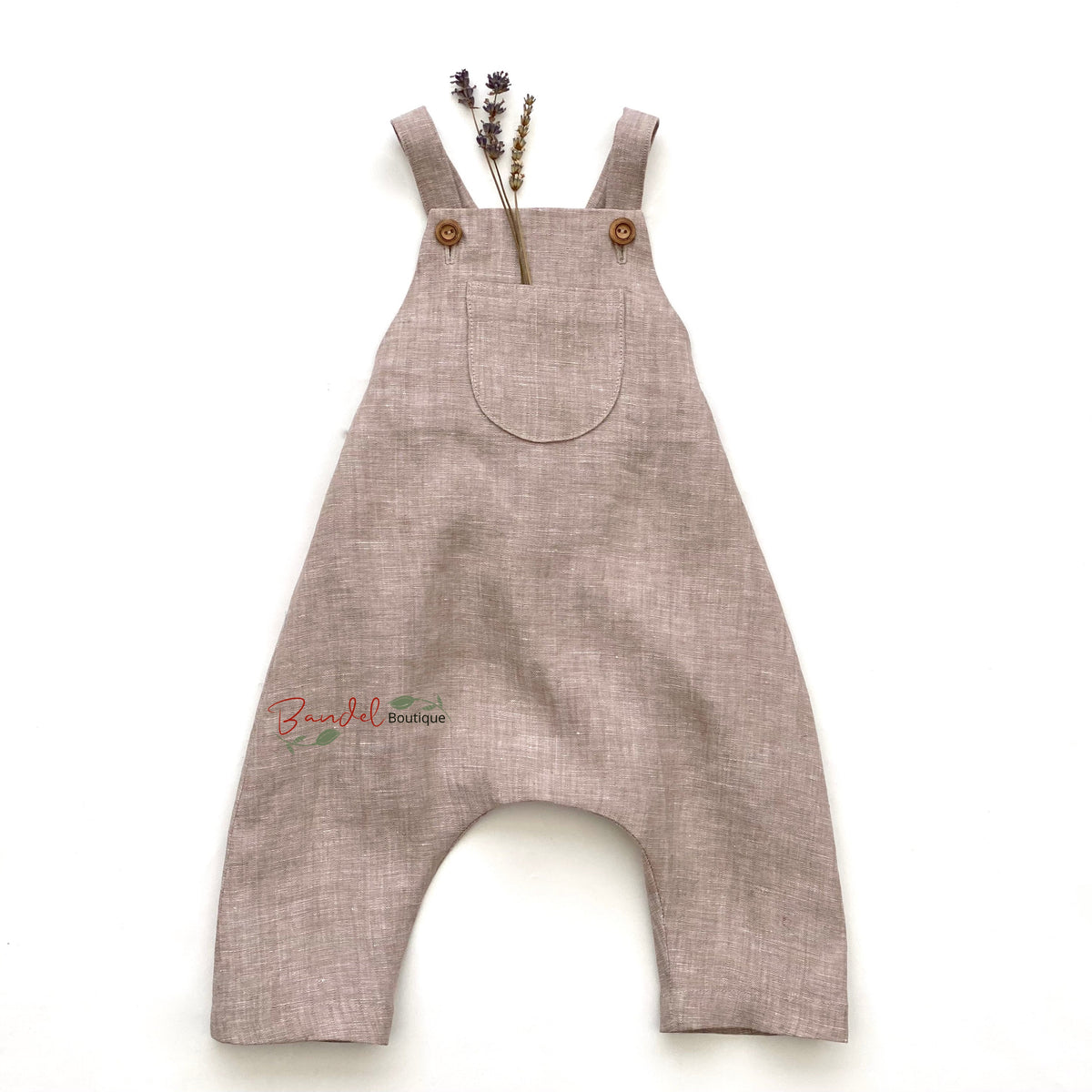 A light brown linen romper with wide legs and shoulder straps, laid flat on a white background with dried lavender stems tucked into the chest pocket.
