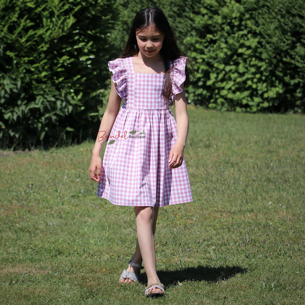 Young girl in a purple checkered dress standing on grass with greenery in the background