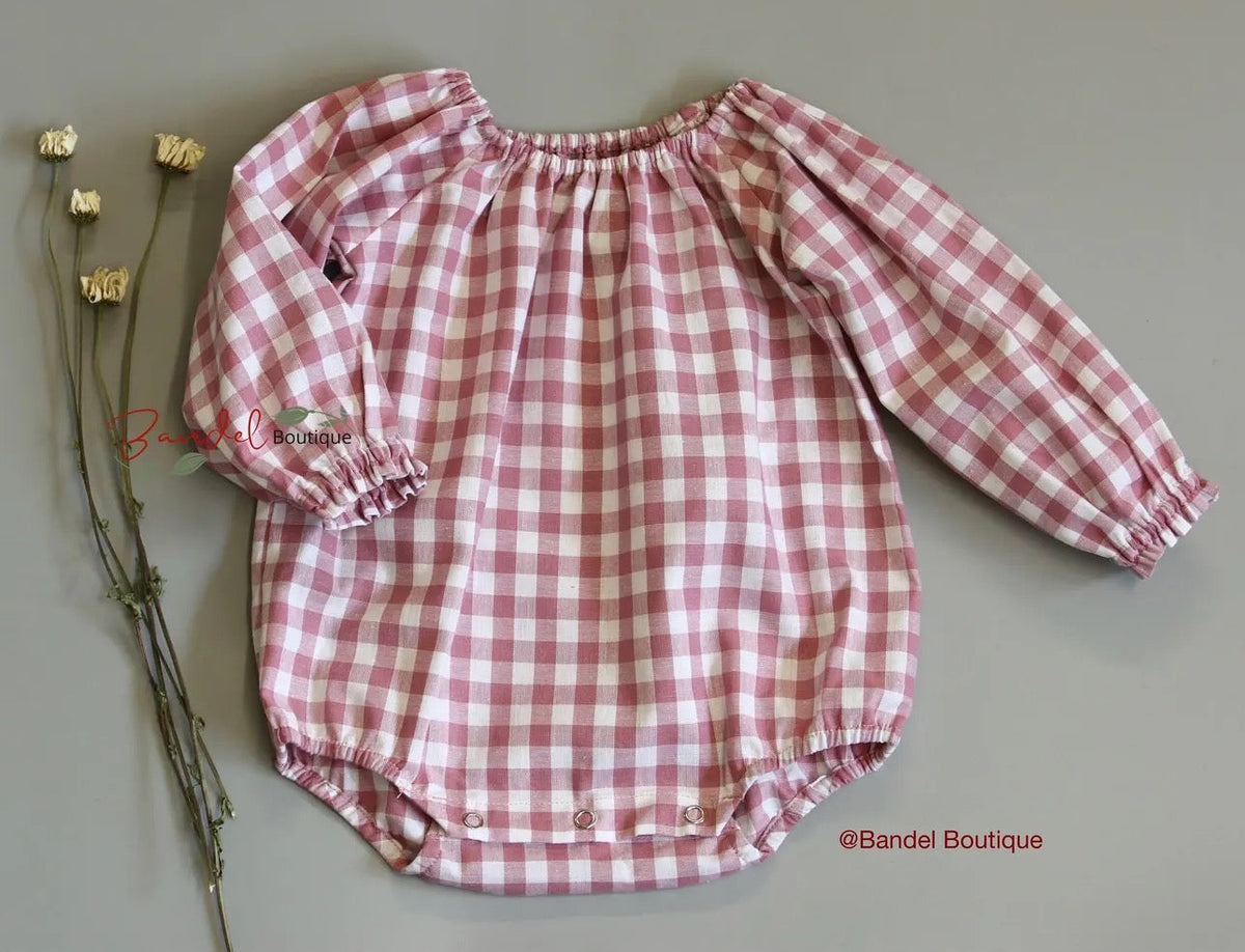 A pink and white gingham long-sleeve baby romper with elasticized cuffs and hem, laid flat on a neutral gray background next to dried yellow flowers. A watermark for 'Bandel Boutique' is visible.