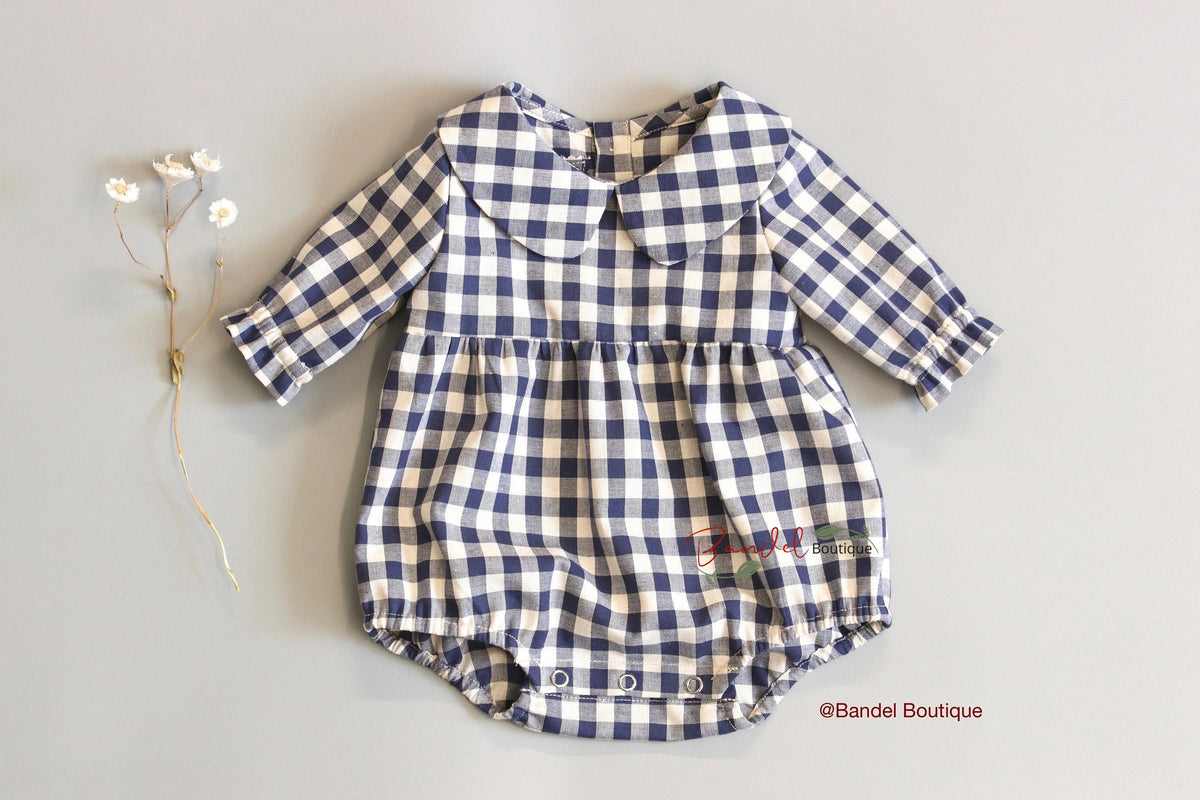 A baby long-sleeve romper in a navy blue and cream gingham check pattern, featuring a Peter Pan collar and snap closures at the crotch, laid flat on a light gray background next to small dried white flowers.