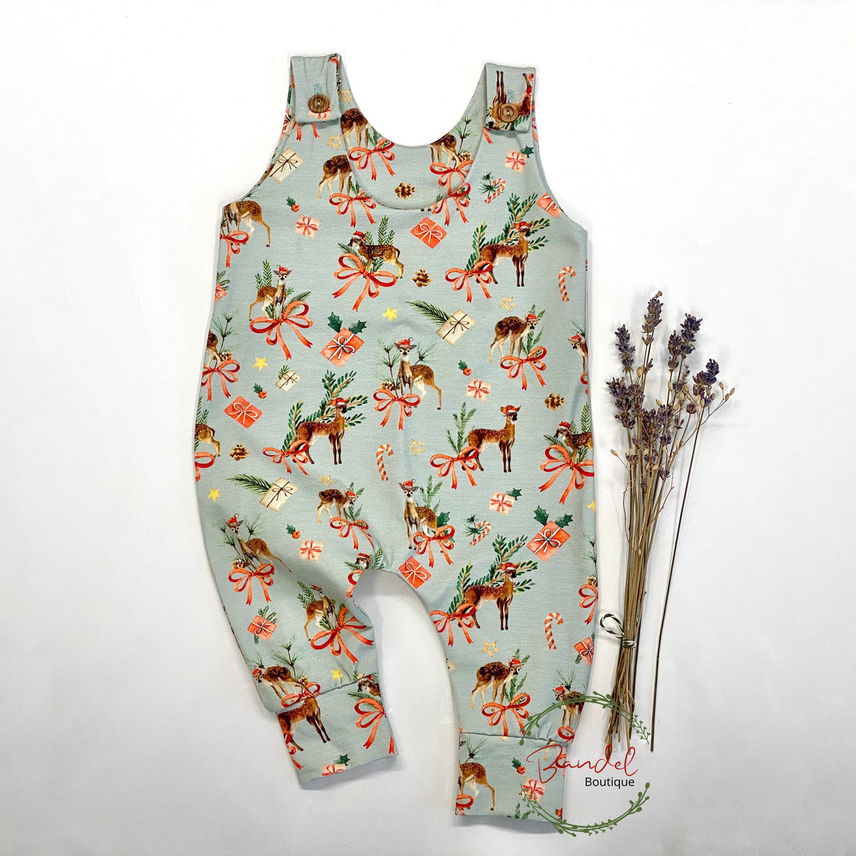 A sleeveless baby romper with a holiday print of deer, red bows, candy canes, and gifts on a sage green background, styled flat-lay with dried lavender on a white background.