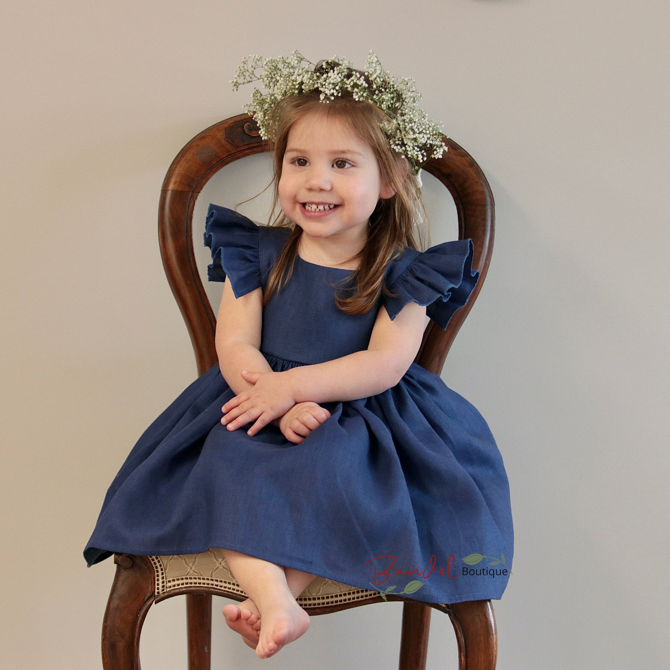 Young girl in a blue dress sitting on a wooden chair with a neutral background