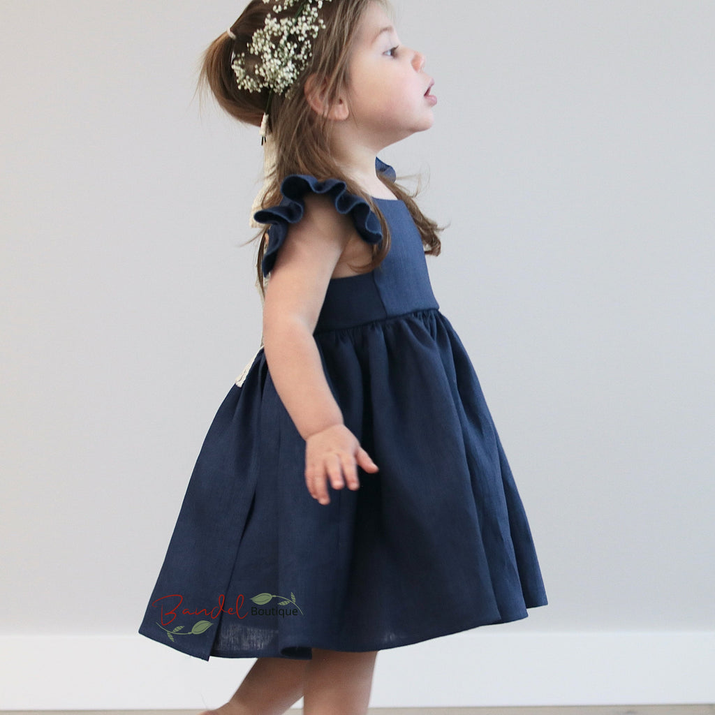 A young girl wearing a royal blue girl's linen dress with a square neckline, flutter sleeves, and a ruffle hem, standing with a decorative headpiece.