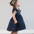 A young girl wearing a royal blue girl's linen dress with a square neckline, flutter sleeves, and a ruffle hem, standing with a decorative headpiece.