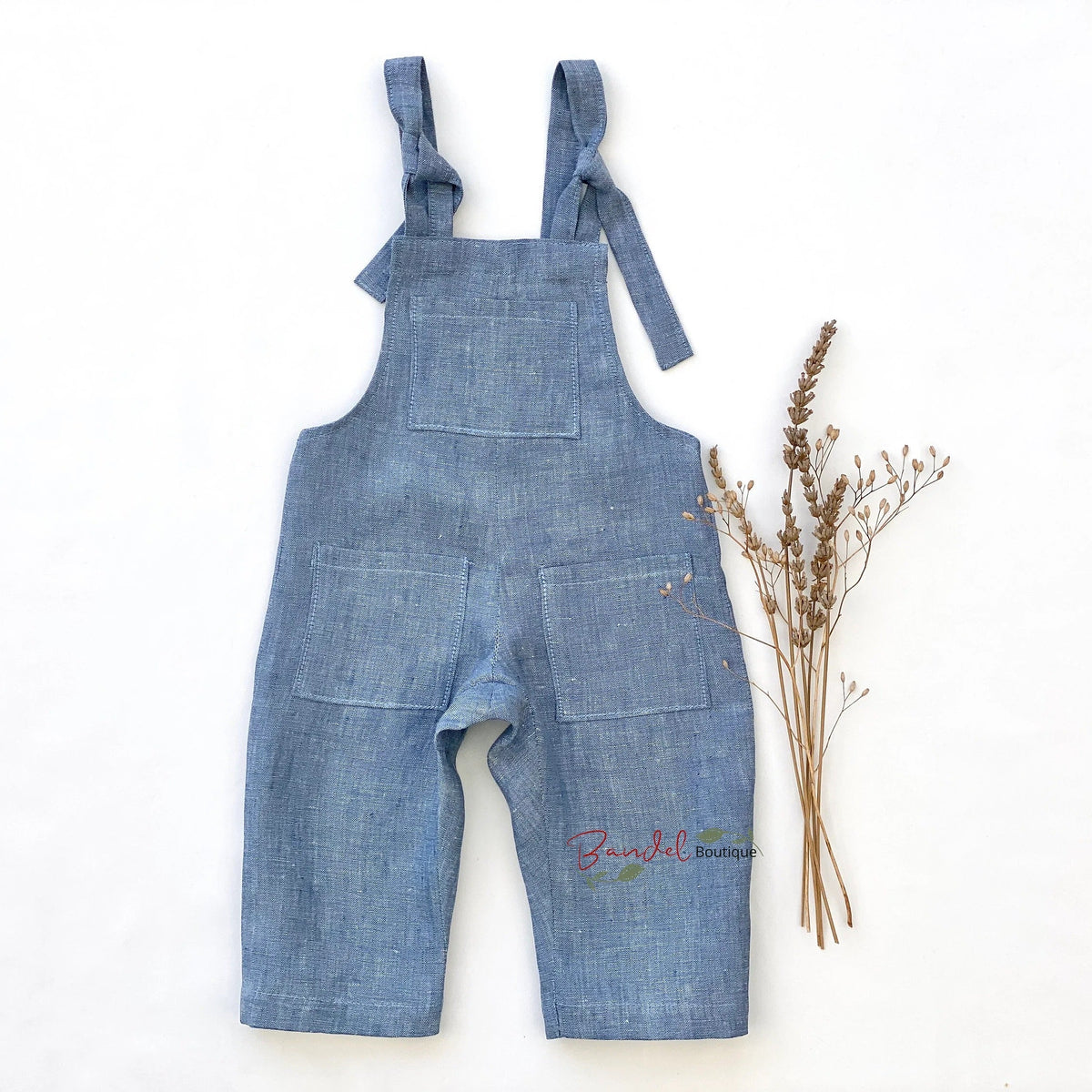 A pair of blue denim baby overalls with tie straps and front pockets, laid flat on a white background next to a small bunch of dried flowers.