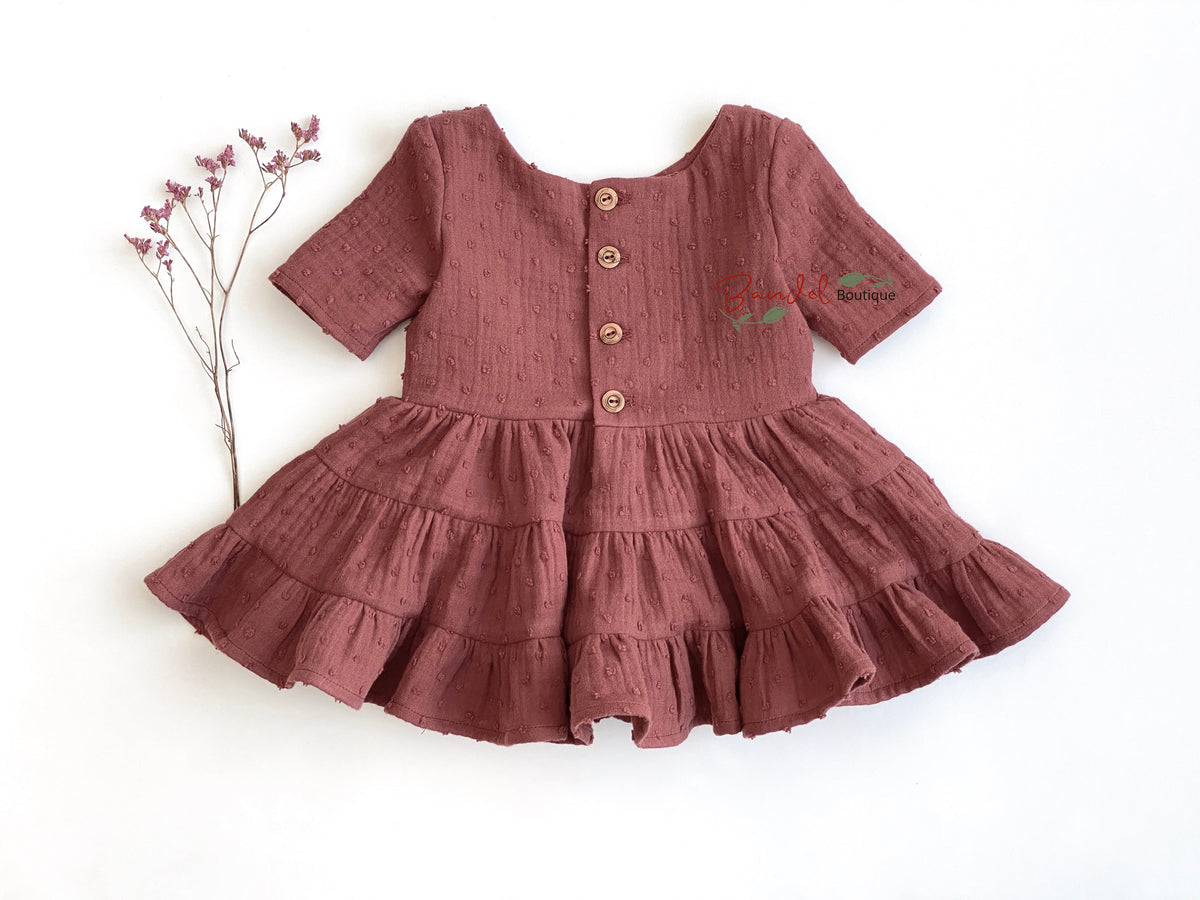 A dusty rose short-sleeve baby dress made of textured linen-like fabric, featuring a tiered ruffled skirt and a button-up front. The dress is laid flat on a white background next to a small sprig of dried pink flowers.