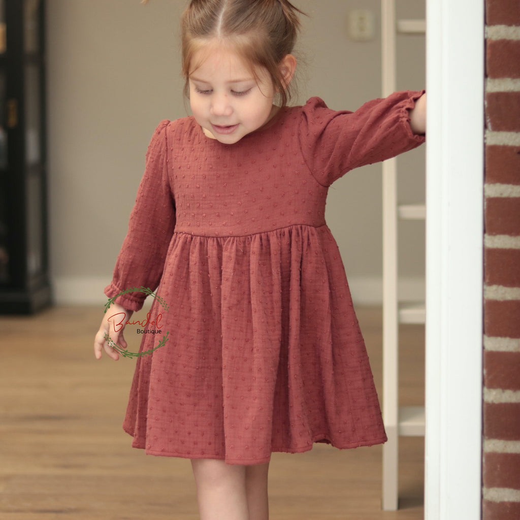 Young girl in a red dress standing in a room with wooden floor and bookshelf.
