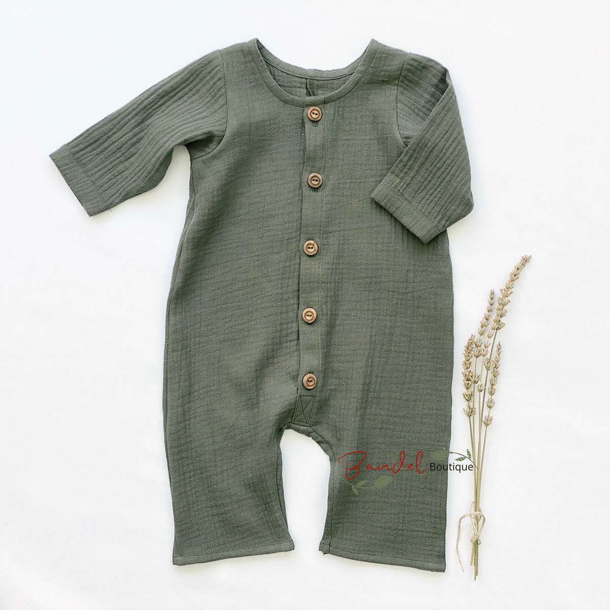 An olive green long-sleeve baby romper with brown wooden buttons, laid flat on a white background next to a small dried sprig of lavender.