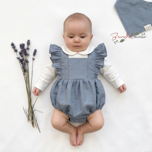 A baby lying on a white surface wearing a dusty blue linen romper with ruffled shoulder straps and a white long-sleeve shirt underneath. A matching dusty blue linen cloth is placed in the top right corner, and a small sprig of lavender is on the left.