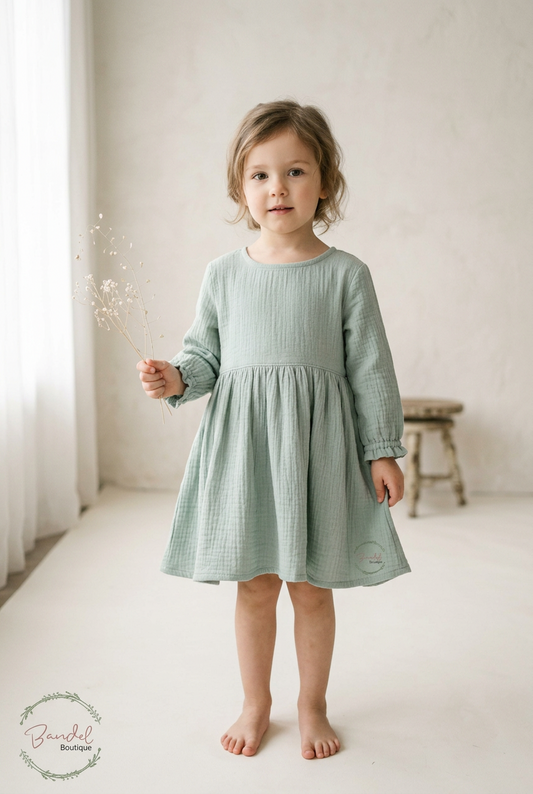 A young child wearing a sage green long-sleeve cotton dress, standing barefoot in a bright, neutral studio setting.