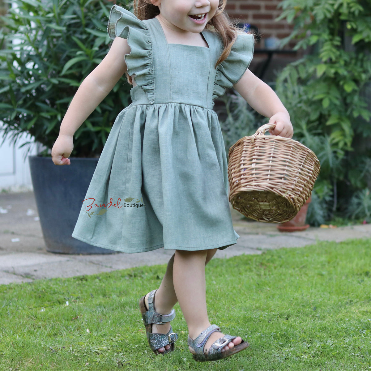 A young girl with long light brown hair is walking on a green lawn in a backyard, wearing a sage green sleeveless dress with ruffled shoulders and carrying a small woven basket.