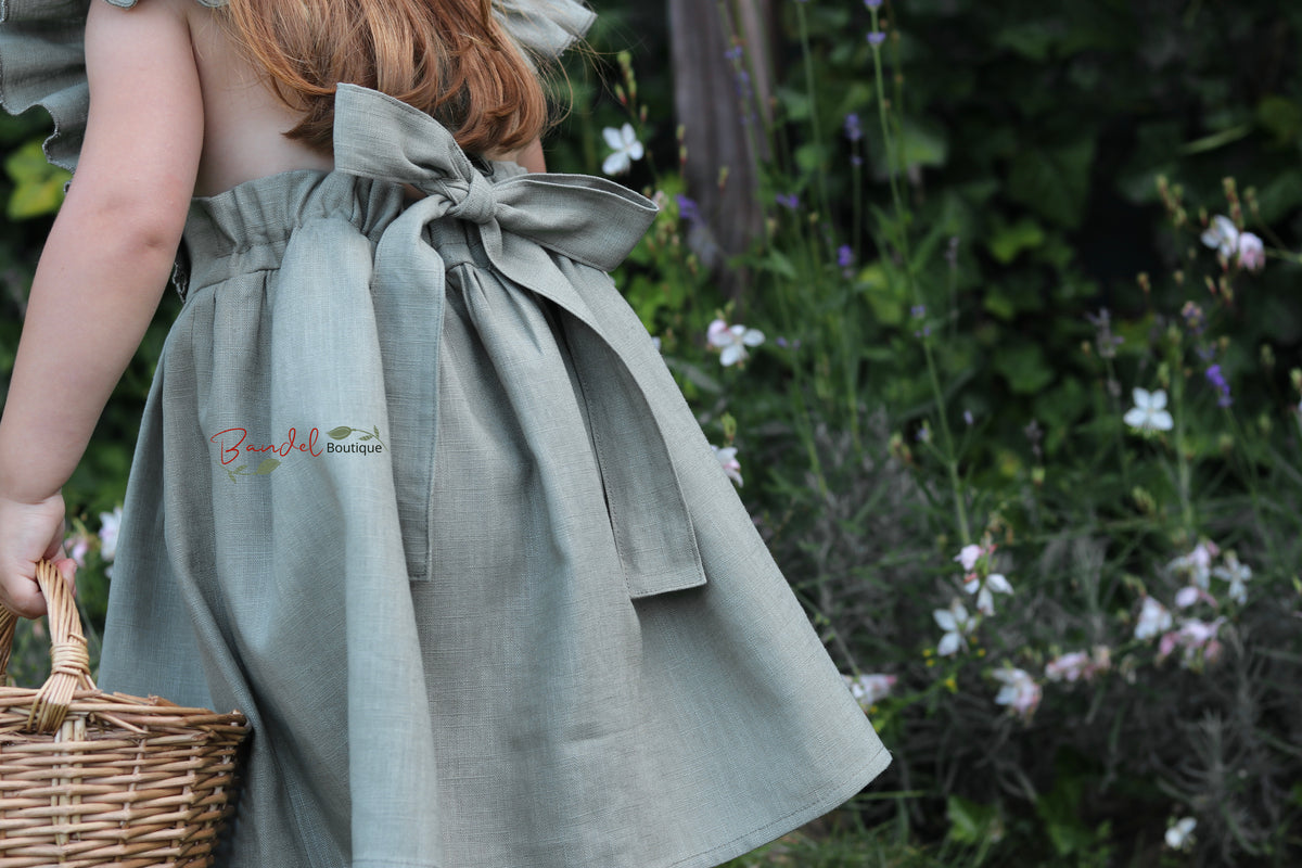 A child wearing a sage green linen dress with a large bow on the back, holding a wicker basket in a garden setting.