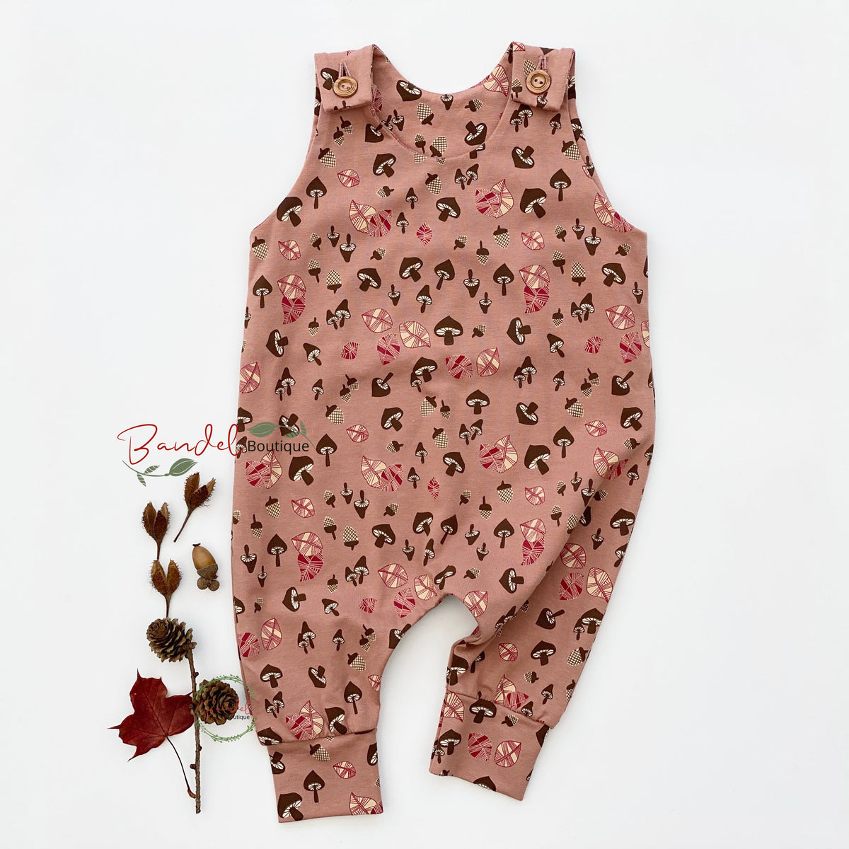 A sleeveless baby romper in dusty rose pink with an all-over print of small brown and pink flowers and faces, laid flat on a white background next to a dried floral stem. A 'Bundel Photography' watermark is visible on the left.