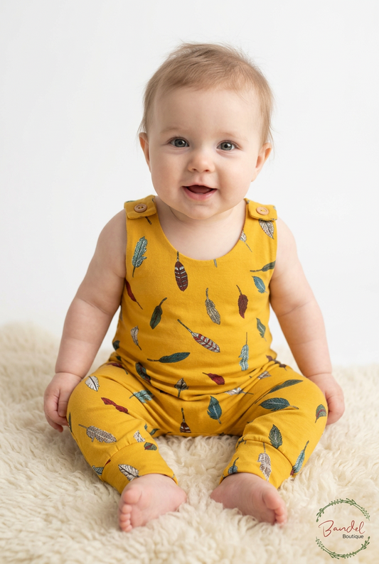 A baby sitting on a cream shag rug wearing a mustard yellow sleeveless romper and matching pants with a colorful feather print. A 'Bundel Boutique' watermark is visible in the bottom right corner.