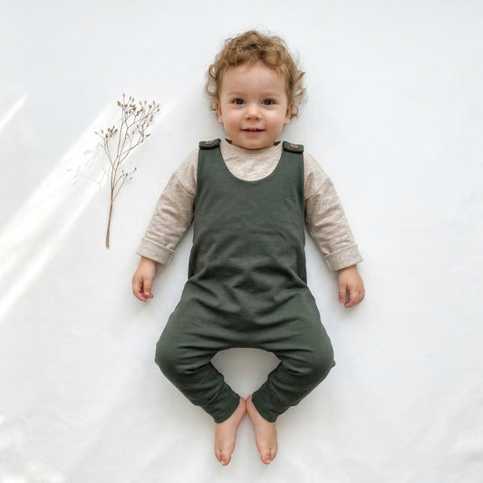 An olive green baby romper with adjustable shoulder straps and small wooden buttons, laid flat on a white background next to a small dried plant stem.