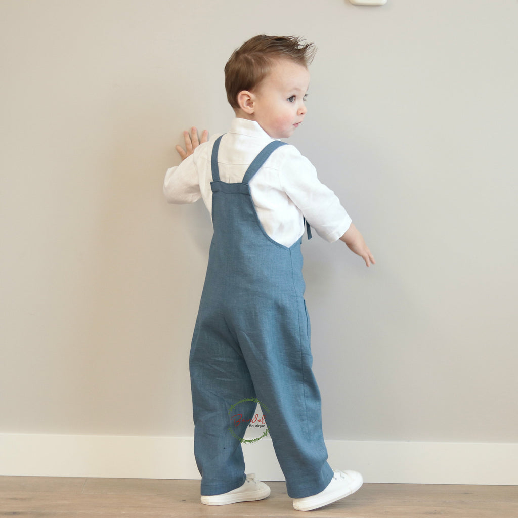 Child wearing blue overalls standing against a plain wall