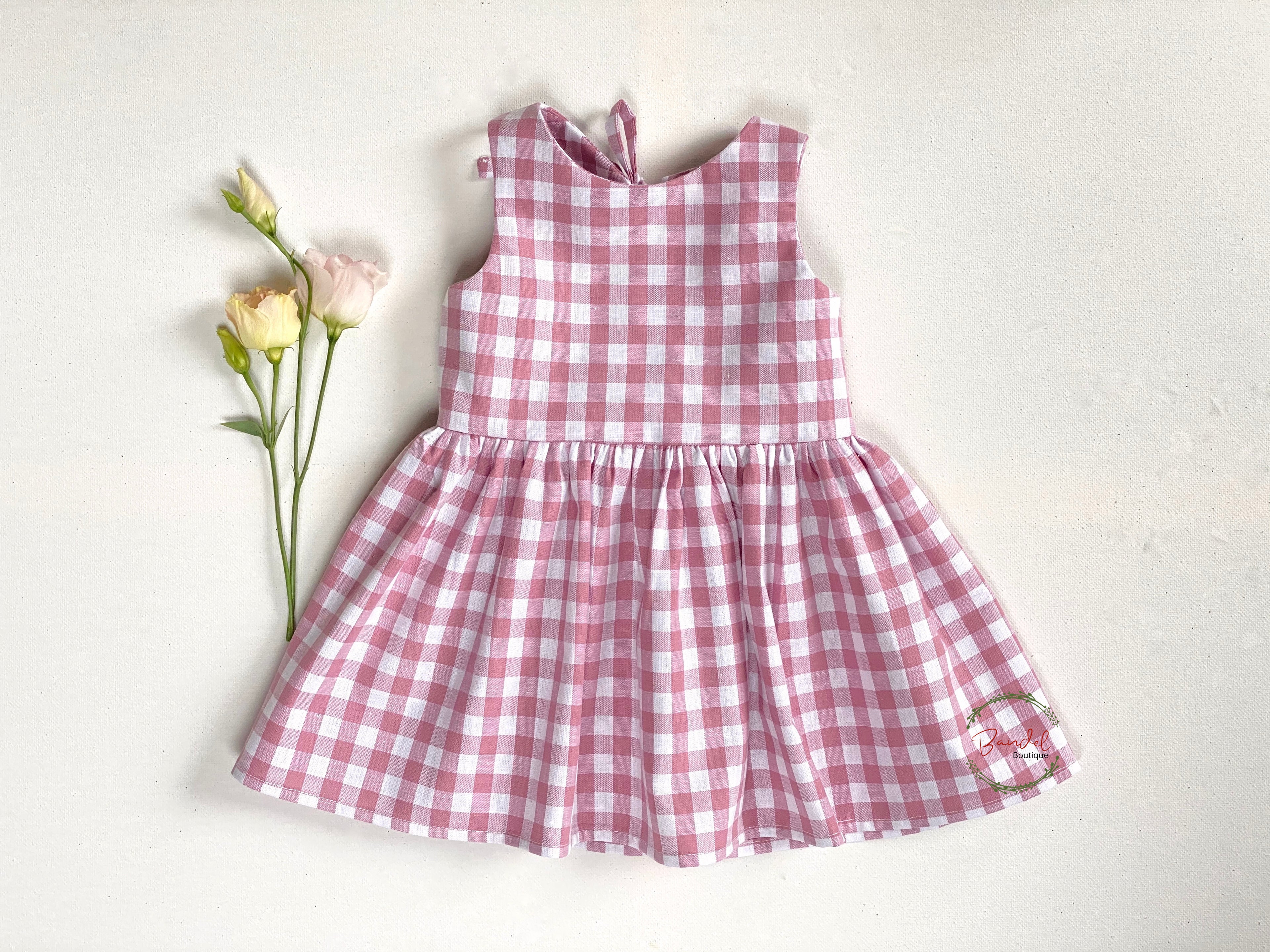 A pink and white checkered cotton dress with a simple front bodice and a large bow-tie, displayed next to a small bouquet of pink roses.