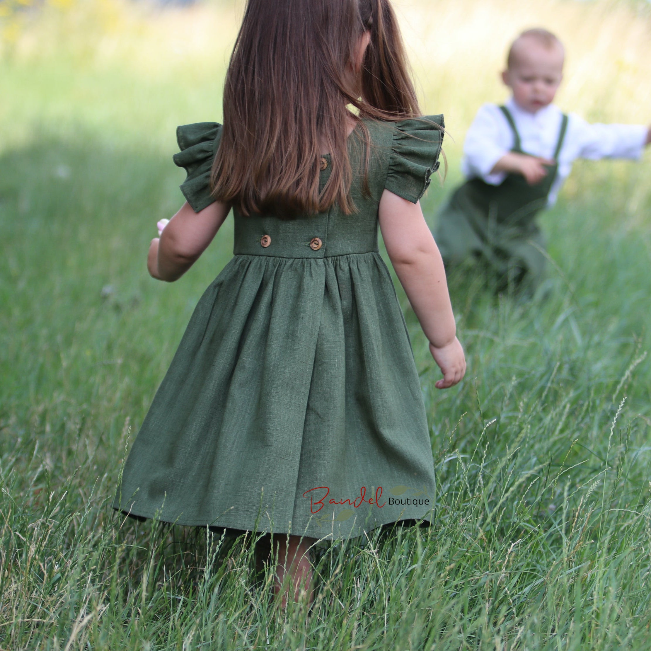 Child in a green dress walking through a grassy field with another child in the background.