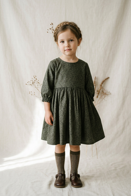 A young girl stands against a draped cream fabric backdrop wearing a dark olive green patterned dress with three-quarter sleeves, matching knee-high socks, and dark brown shoes.