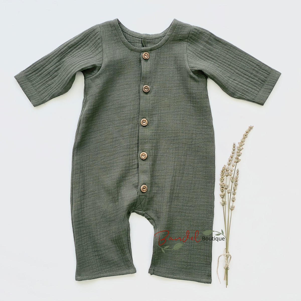 An olive green ribbed cotton muslin baby romper with brown wooden buttons, laid flat on a white background next to a small sprig of dried wheat.
