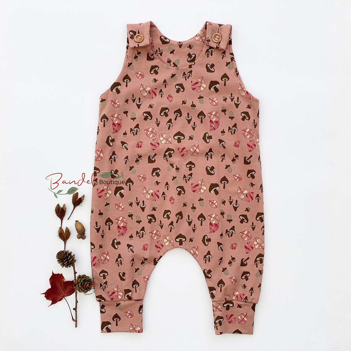 A baby romper in dusty rose pink with an all-over print of small brown and pink mushrooms, laid flat on a white background next to a small dried botanical sprig. The romper has shoulder straps with wooden buttons and cuffed ankles.