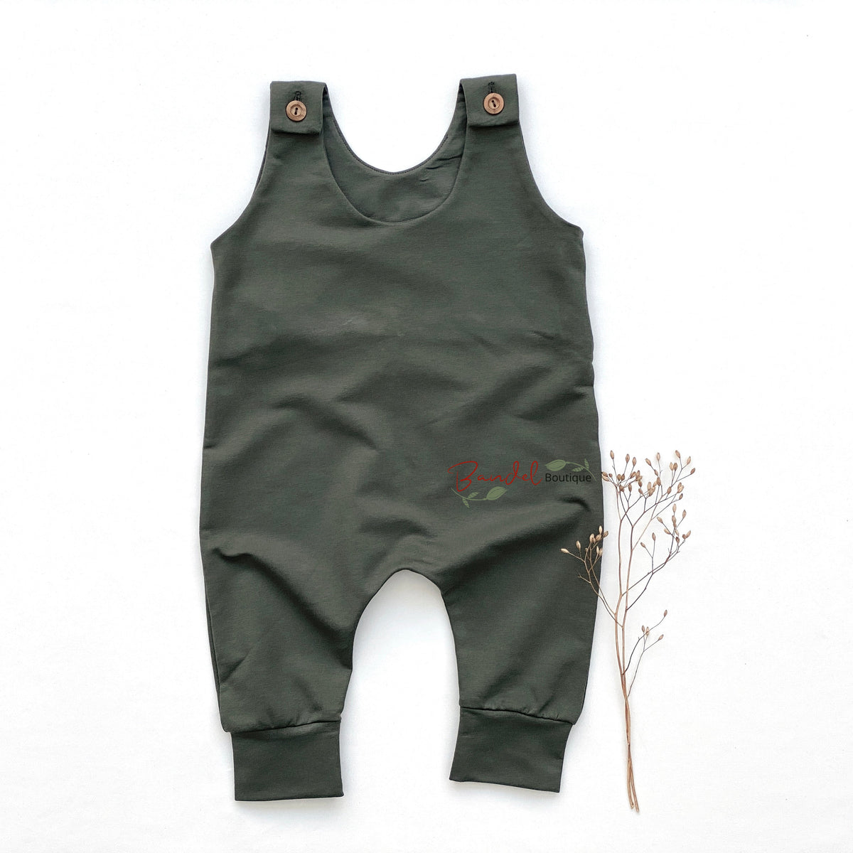 An olive green baby romper with adjustable shoulder straps and small wooden buttons, laid flat on a white background next to a small dried plant stem.